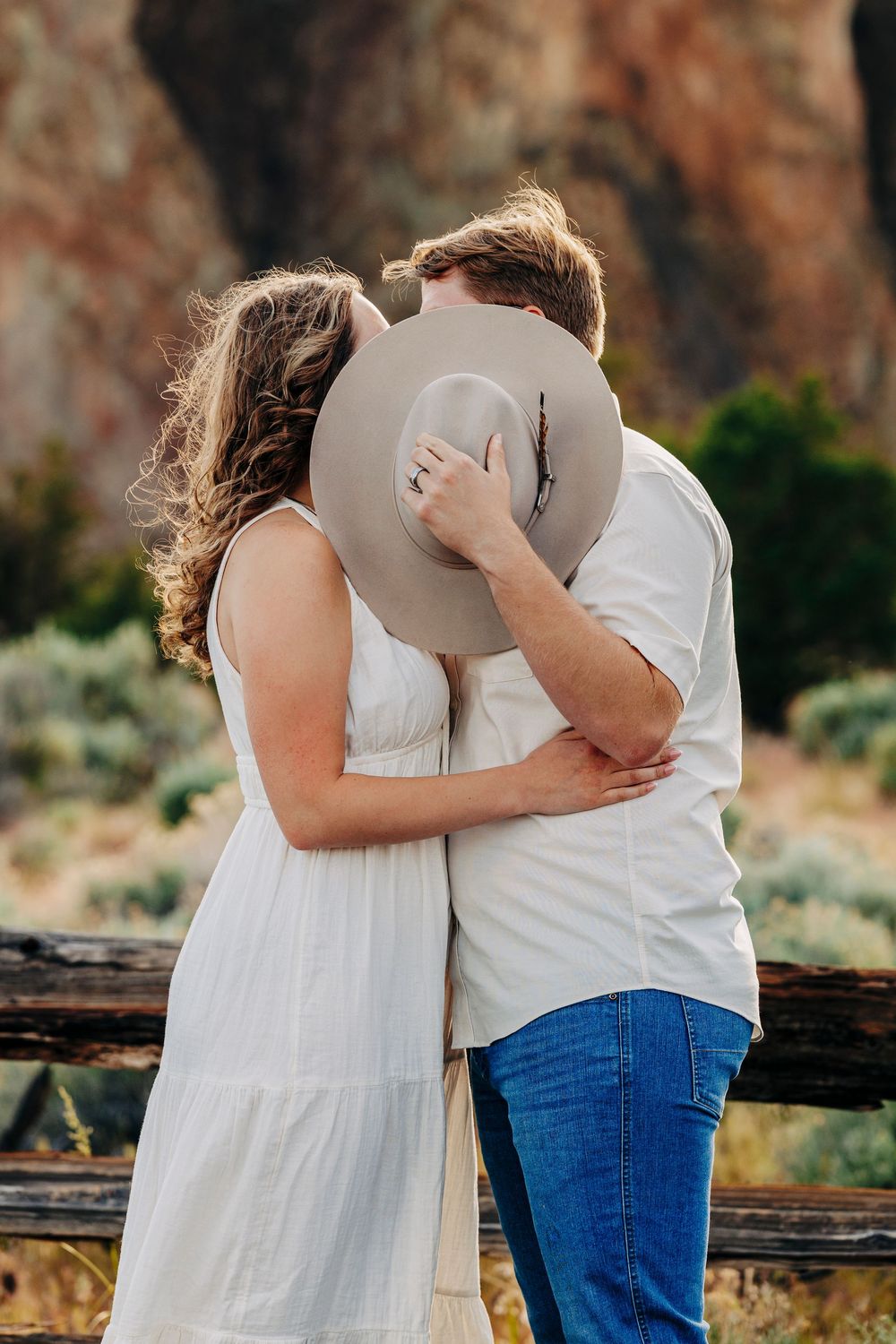 A romantic couple embraces at sunset against a backdrop of red sandstone cliffs in a scenic desert landscape.