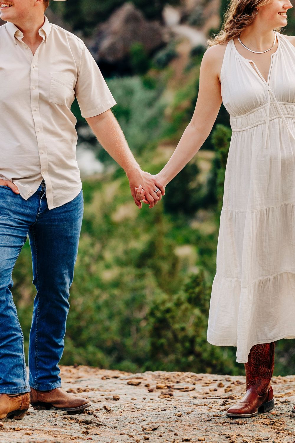 A couple in western attire holding hands while standing on a dirt path outdoors.