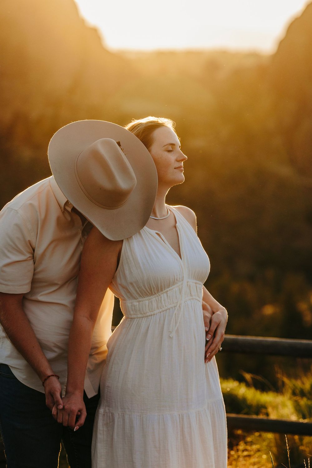 A couple in western wear embraces during golden hour with scenic mountain backdrop.