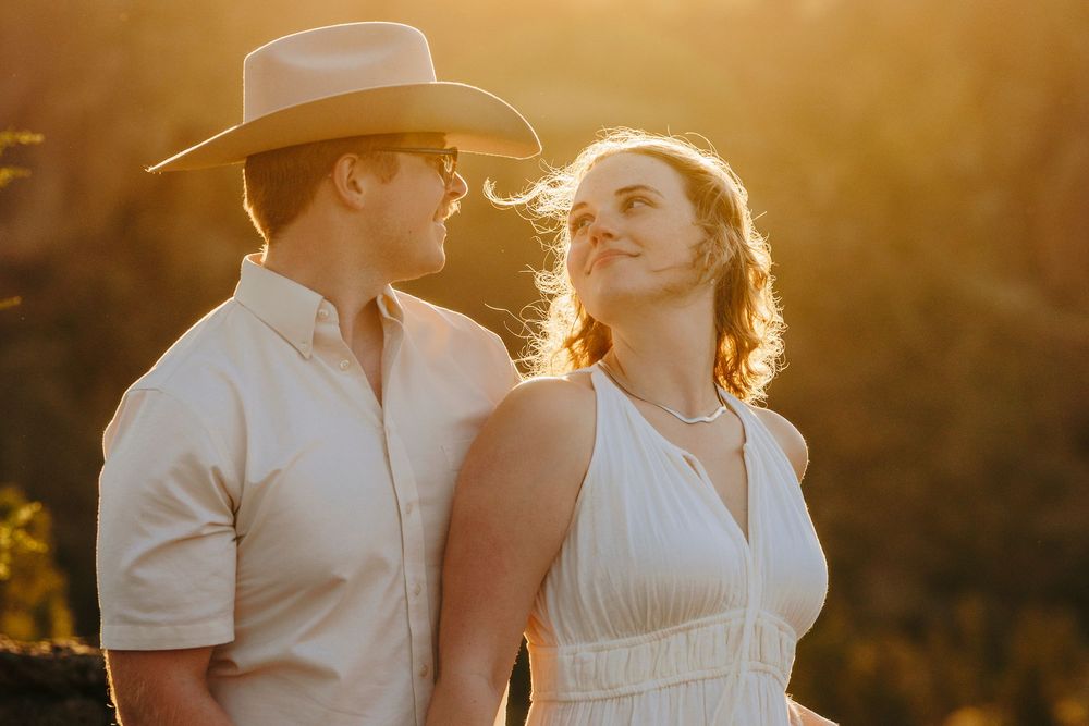 A romantic couple poses together at golden hour, with one wearing a cowboy hat and white summer attire.