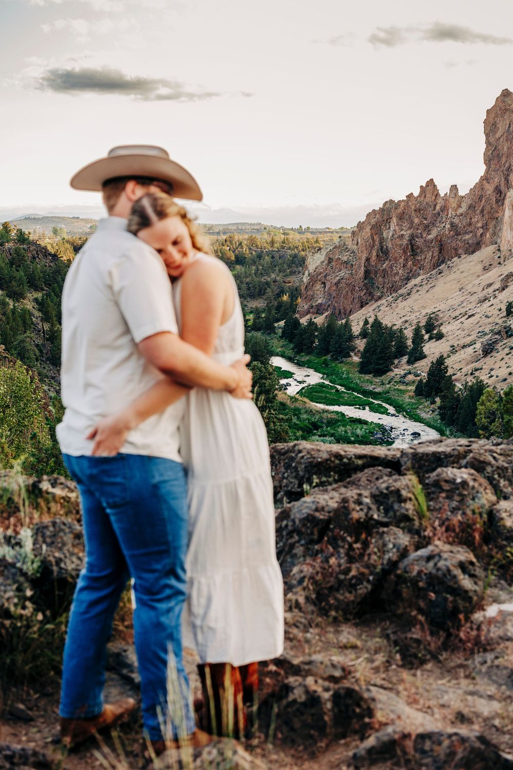 Couple stands together on scenic overlook with dramatic canyon rock formations and winding river below.