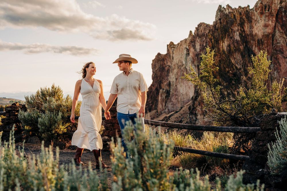 A couple walks hand in hand on a desert trail with dramatic rock formations in the background.