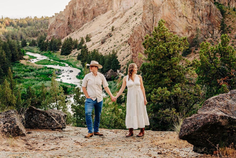 A couple holds hands while standing on a rocky overlook with a scenic canyon valley below.