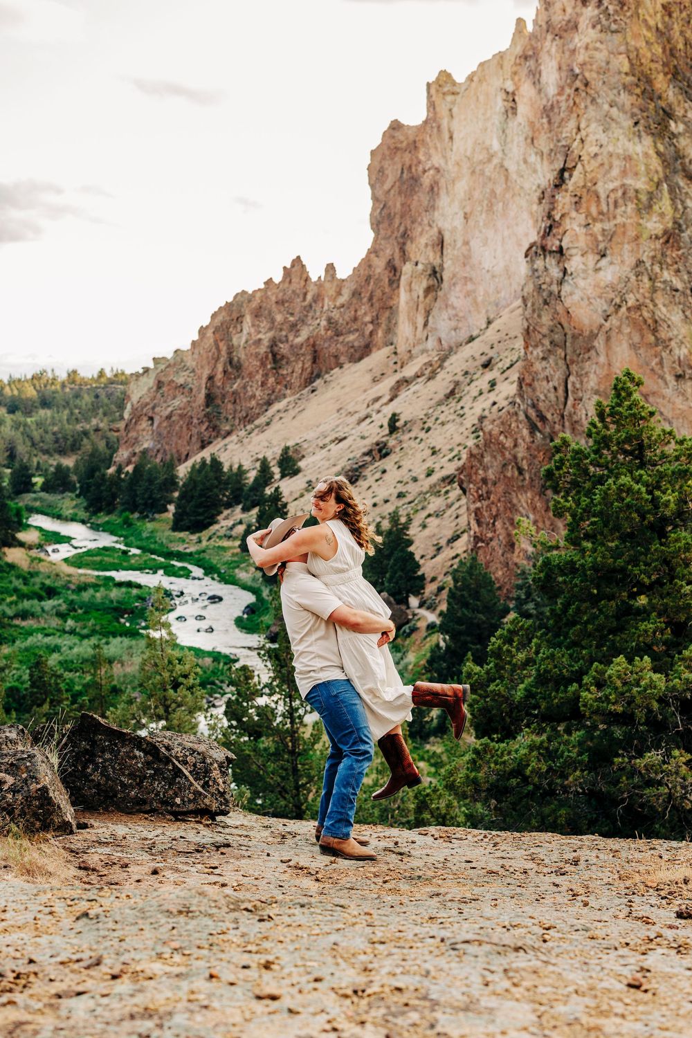 Two people share a romantic embrace against the backdrop of dramatic rock cliffs and a winding river at Smith Rock State Park.