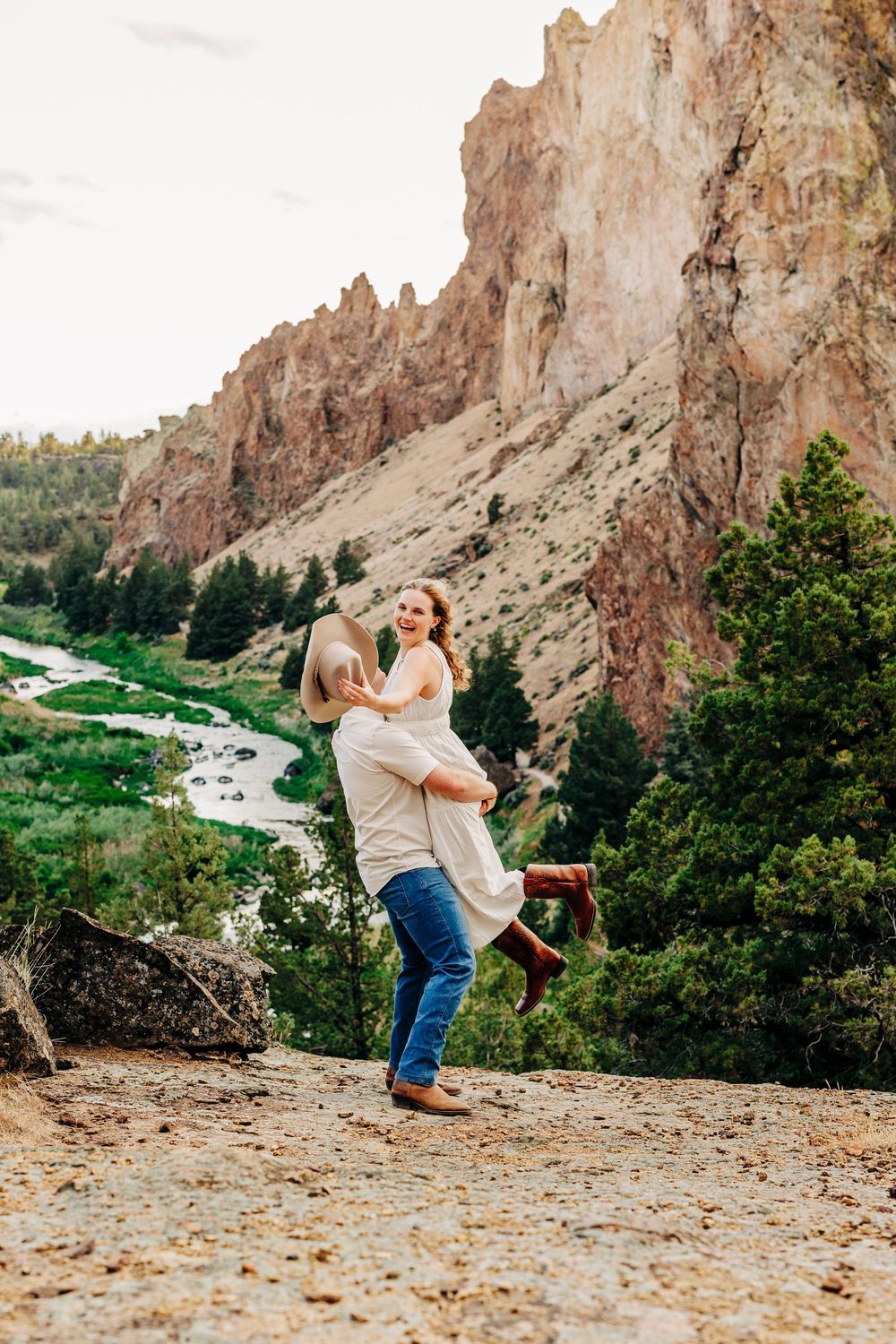 A couple embraces in front of dramatic rocky cliffs with winding road below in nature setting.