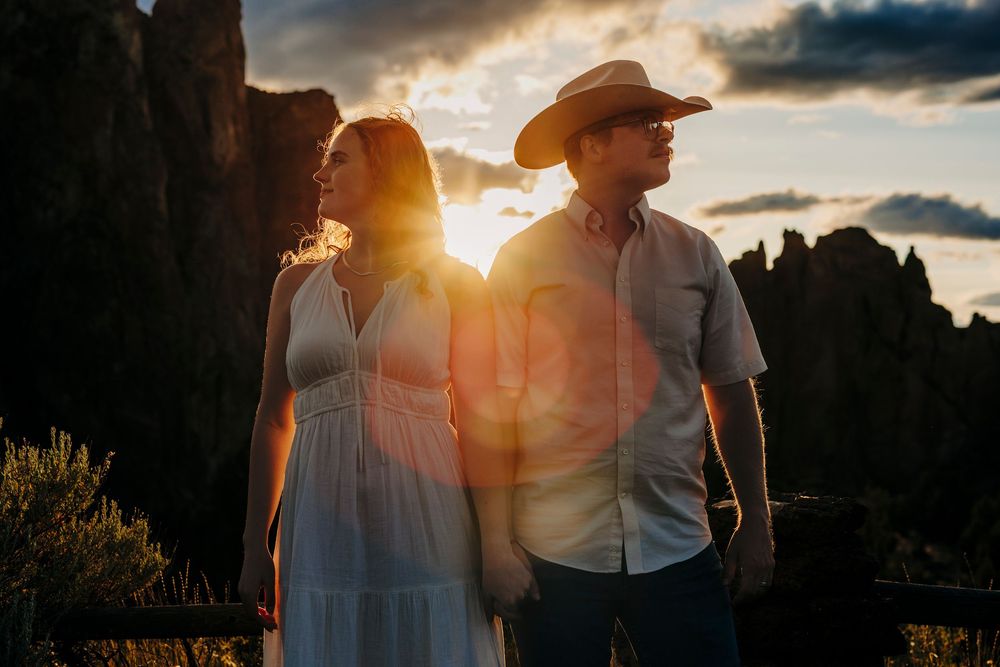 Silhouetted couple stands together against dramatic sunset and rocky mountain backdrop in desert landscape.