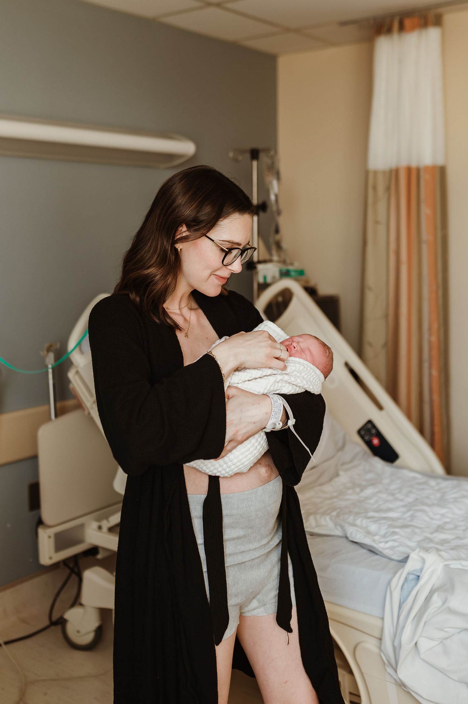 mom in her robe and post party underwear holding her newborn baby in the hospital room