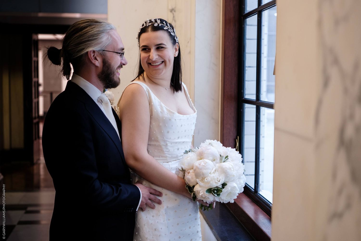 A bride in a white lace dress holds a bouquet of white flowers while standing near a window.