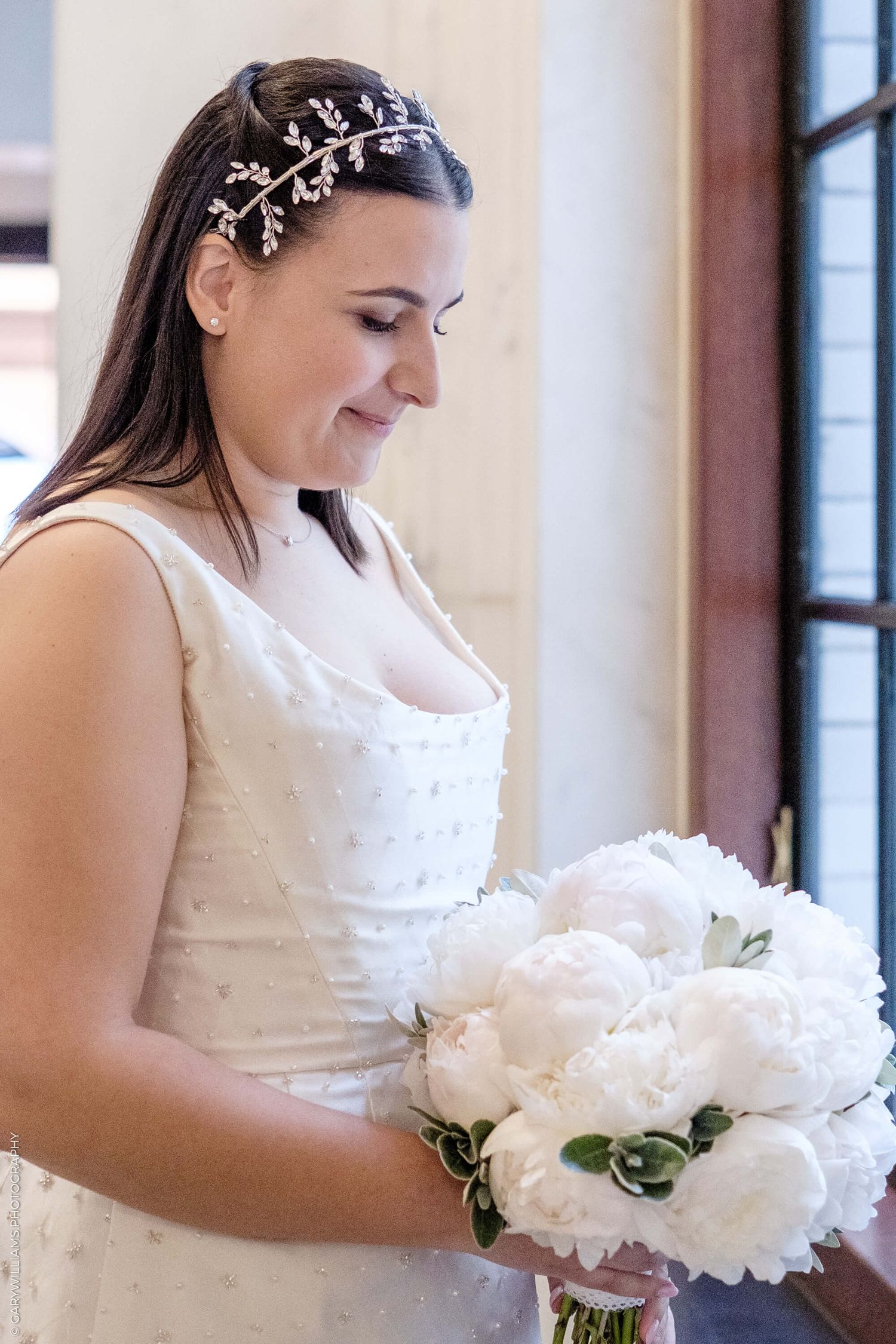 Elegant bride in white dress with delicate crystal hairpiece holding a bouquet of white peonies by window.