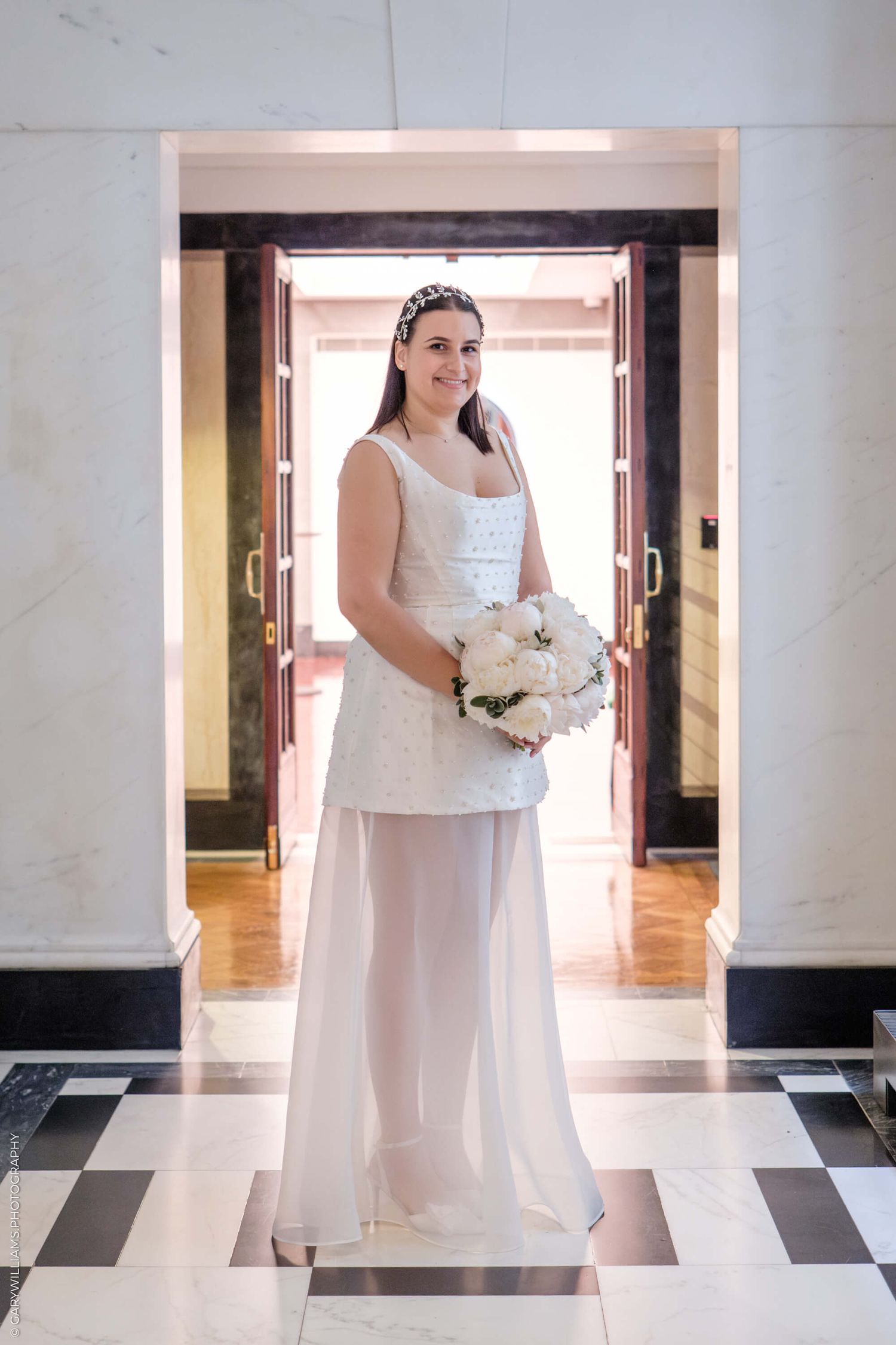 A bride in a white dress stands gracefully in an elegant lobby interior with marble tile flooring and columns.