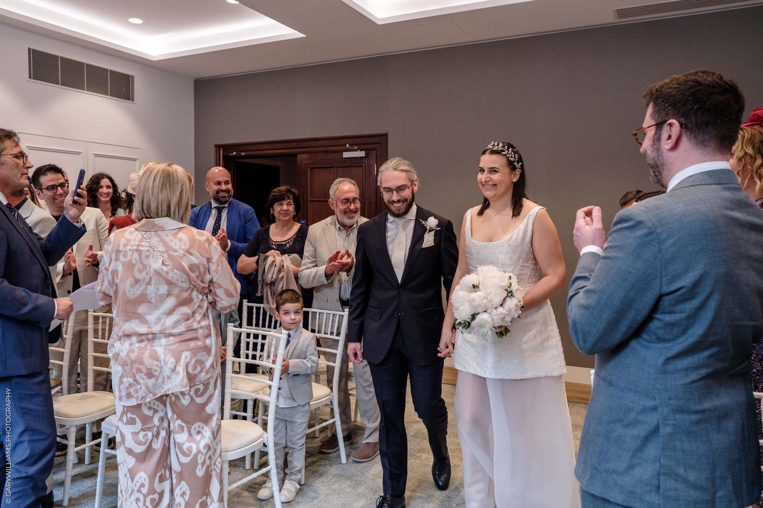 Wedding guests applaud and smile during an intimate indoor ceremony at a modern venue with white chiavari chairs.