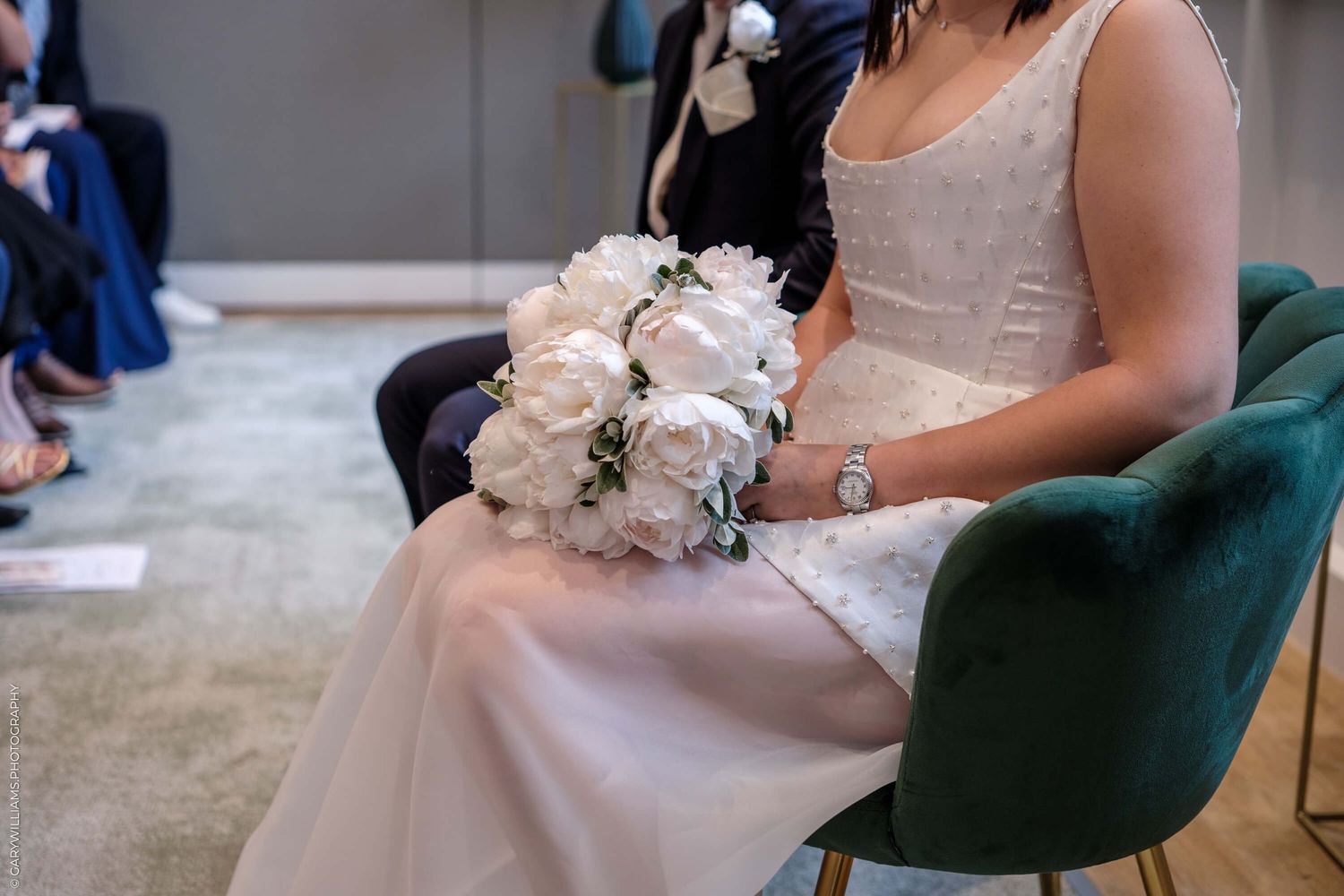 Someone in a light-colored formal dress sits in an emerald green chair holding a delicate bouquet of pale pink flowers.