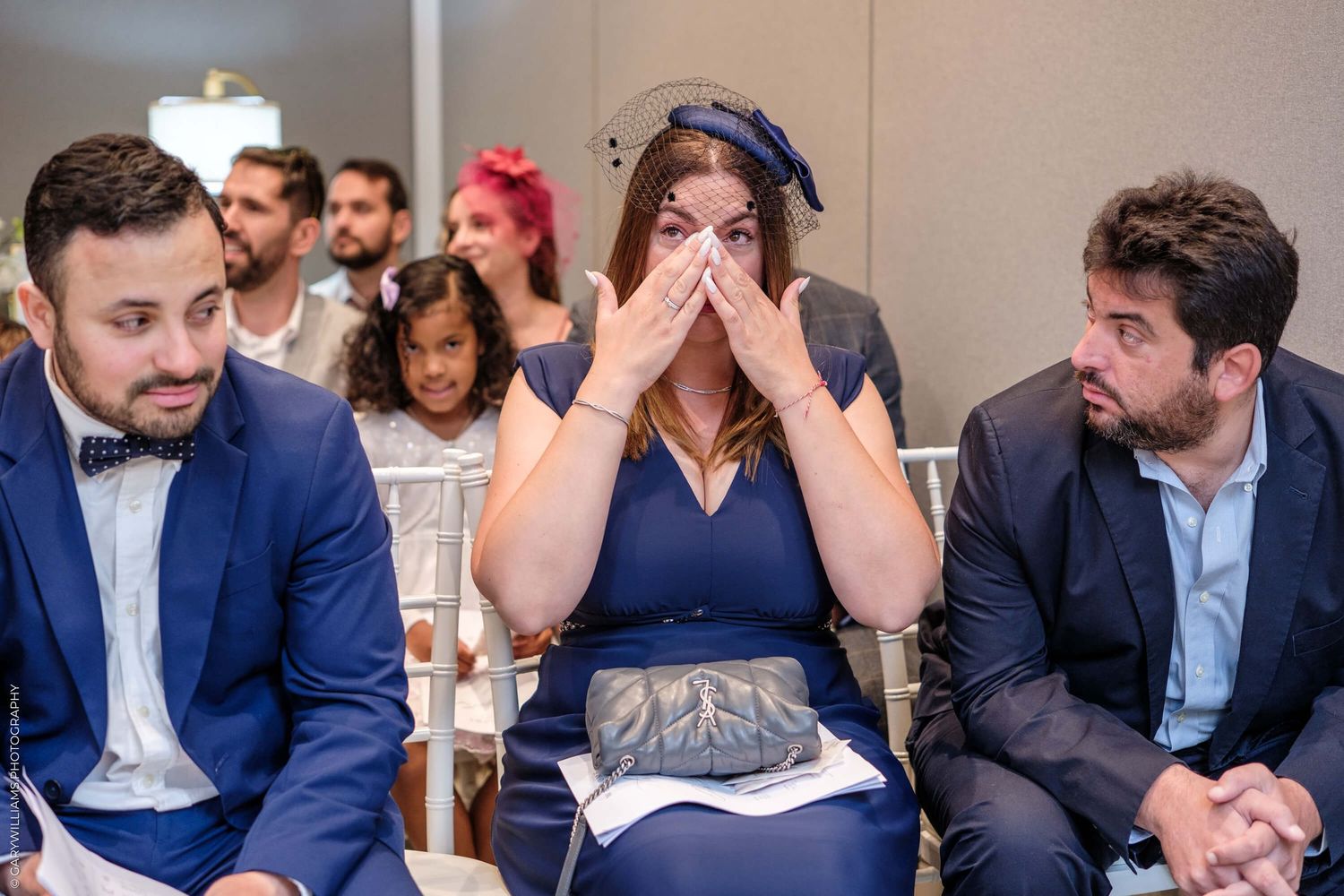 Wedding guests react emotionally during a ceremony while seated in rows of white chairs.