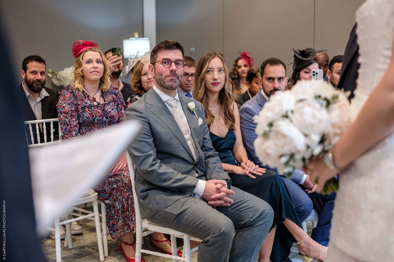 Guests are seated in white chairs during an indoor wedding celebration with sunlit decor.