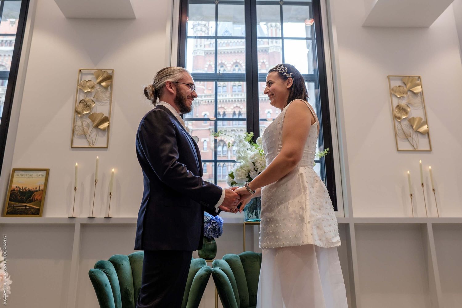 A couple shares an intimate moment during their wedding ceremony in an elegant room with tall windows and gold artwork.