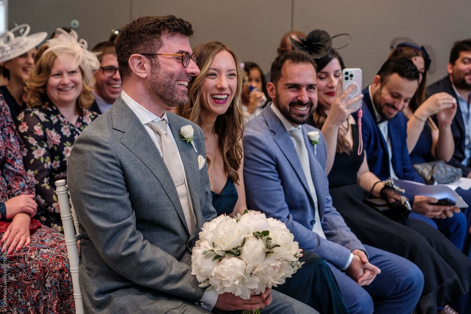 Wedding guests sitting in rows smile and laugh during a joyful indoor ceremony.
