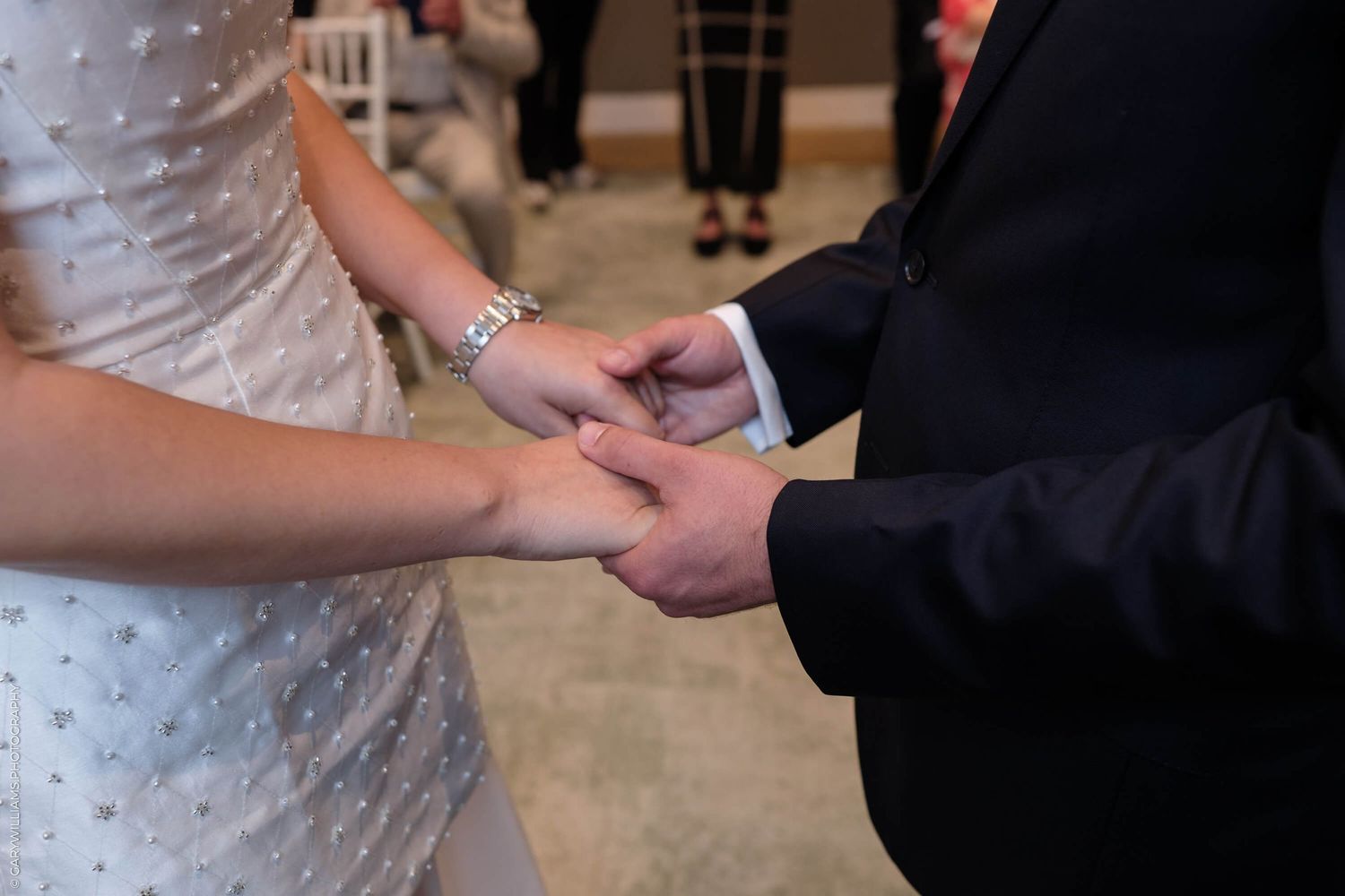 Close-up of newlyweds holding hands during wedding ceremony showing ring and lace dress details.