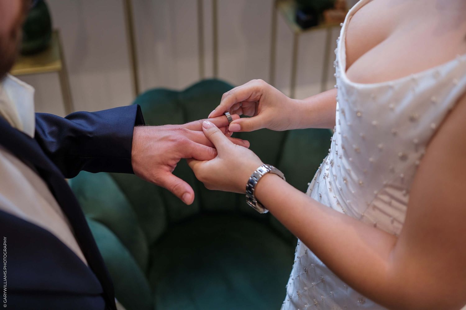 Close-up shot of hands exchanging wedding rings during an intimate ceremony against a dark green background.