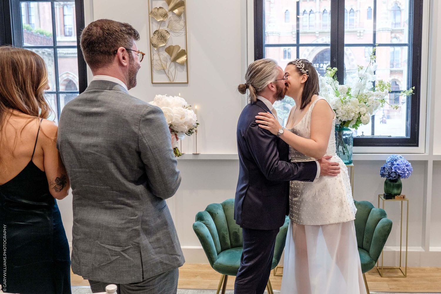 Wedding guests capture the moment as newlyweds share their first kiss in an elegant room with emerald velvet chairs.
