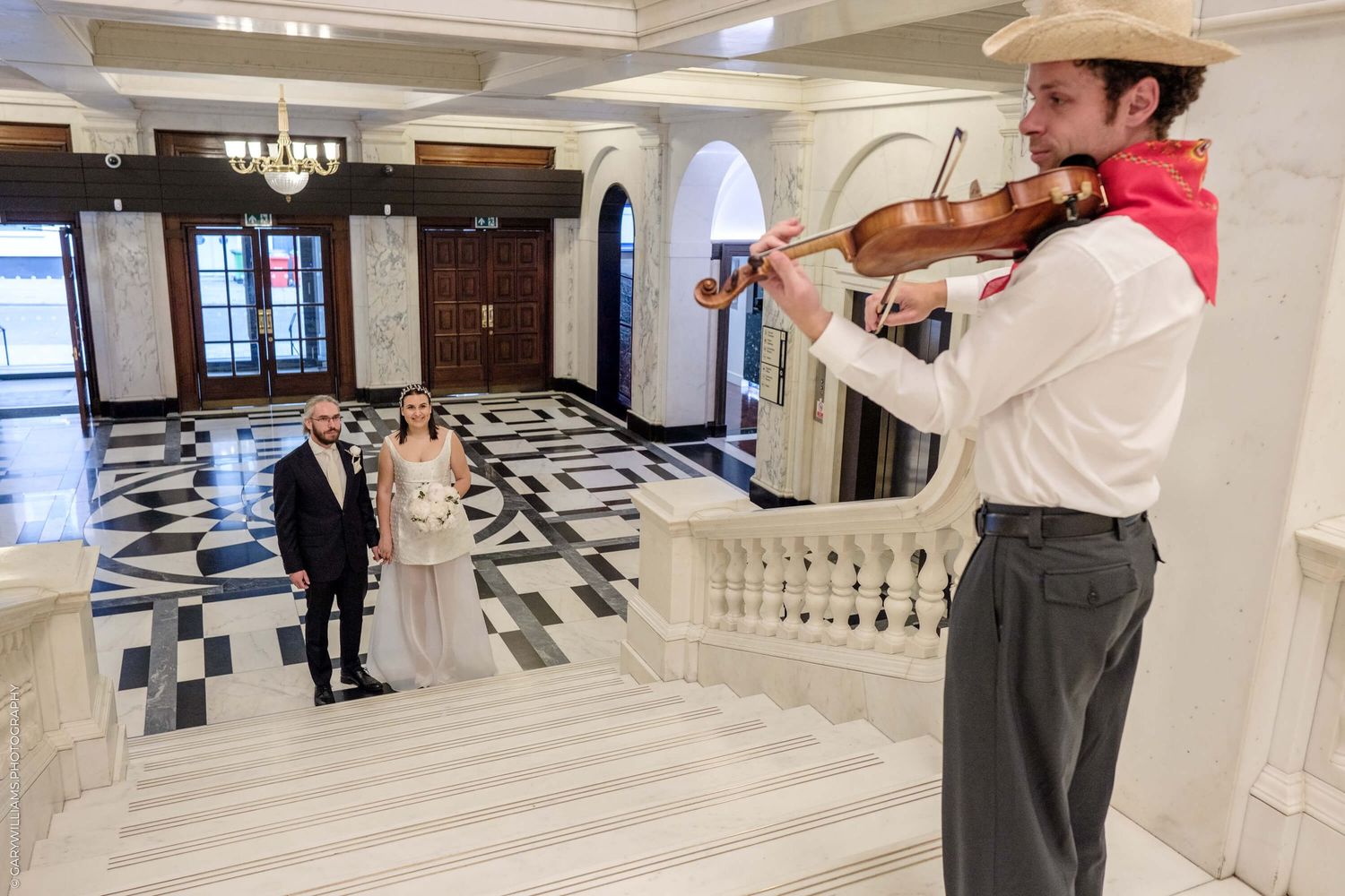A violinist in cowboy attire performs in a grand marble-floored lobby with checkered tiles and elegant decor.