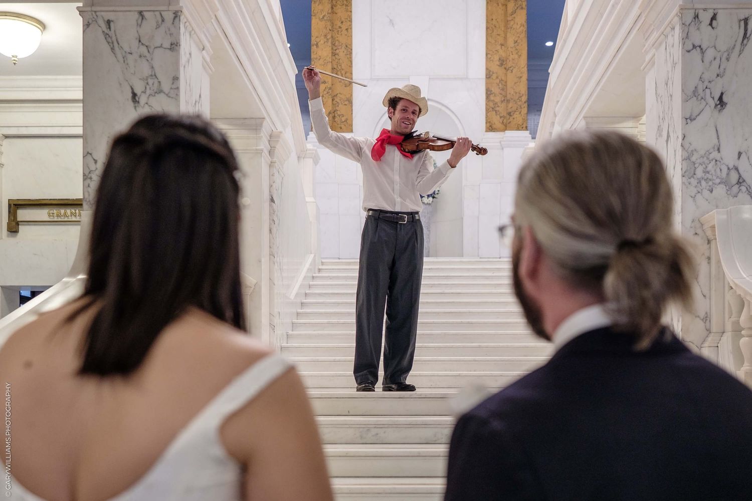 Musician in white shirt and gray pants performs violin solo on ornate marble staircase while onlookers watch from below.