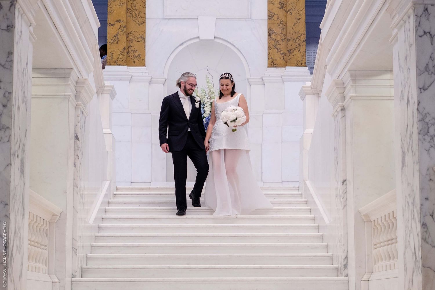Elegant couple stands on grand white marble staircase in formal wedding attire during ceremony.