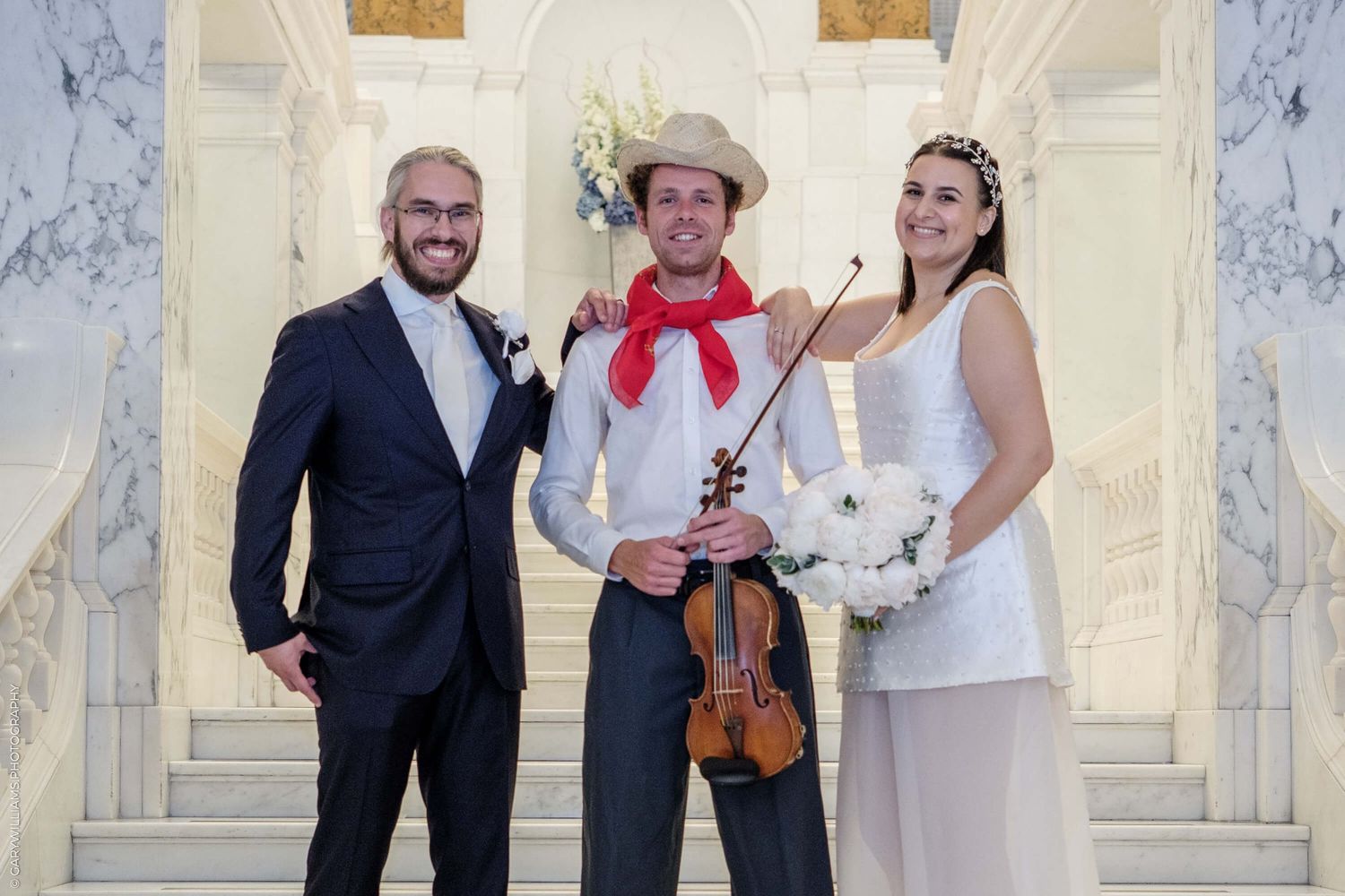 Three people dressed formally stand together at a marble staircase, with one holding a violin and wearing a red bow tie.