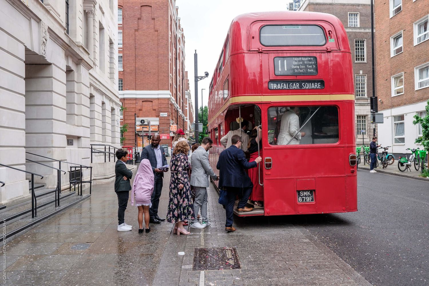 Groups of people walk near iconic red London buses parked along a city street on a cloudy day.