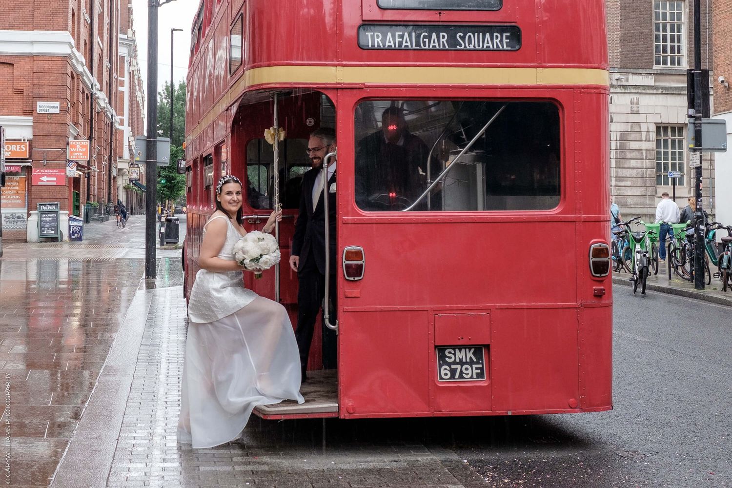 Someone in a white dress boards a vintage red London double-decker bus on a rainy city street.