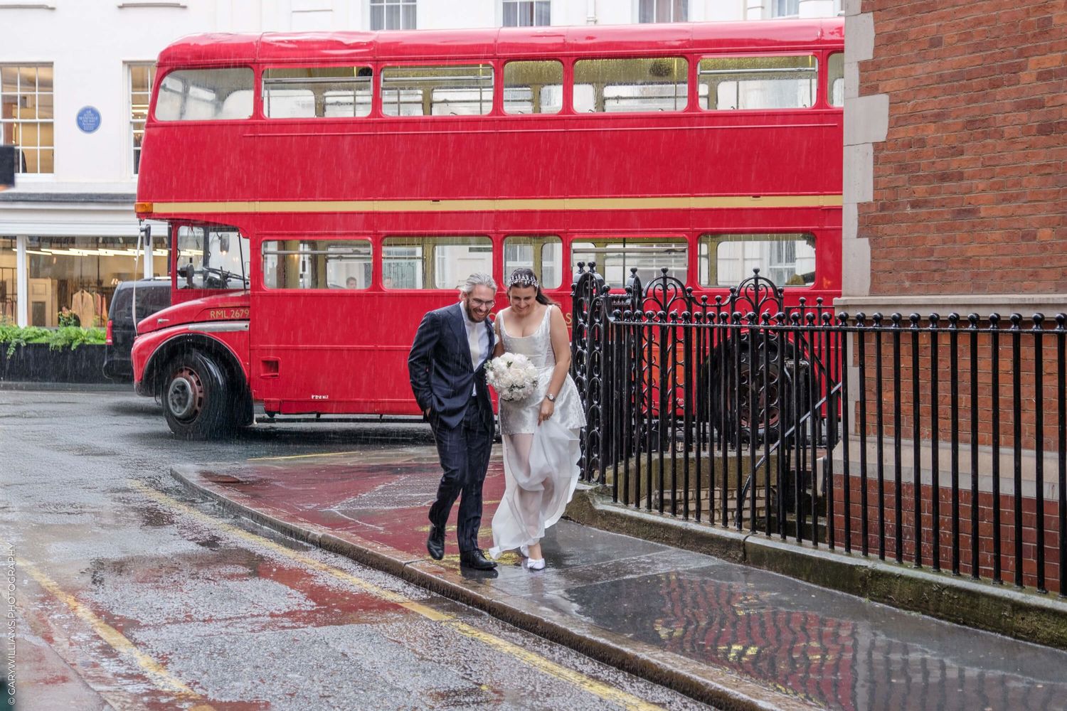 A vintage red double-decker bus parked on a rainy street in front of a brick building.
