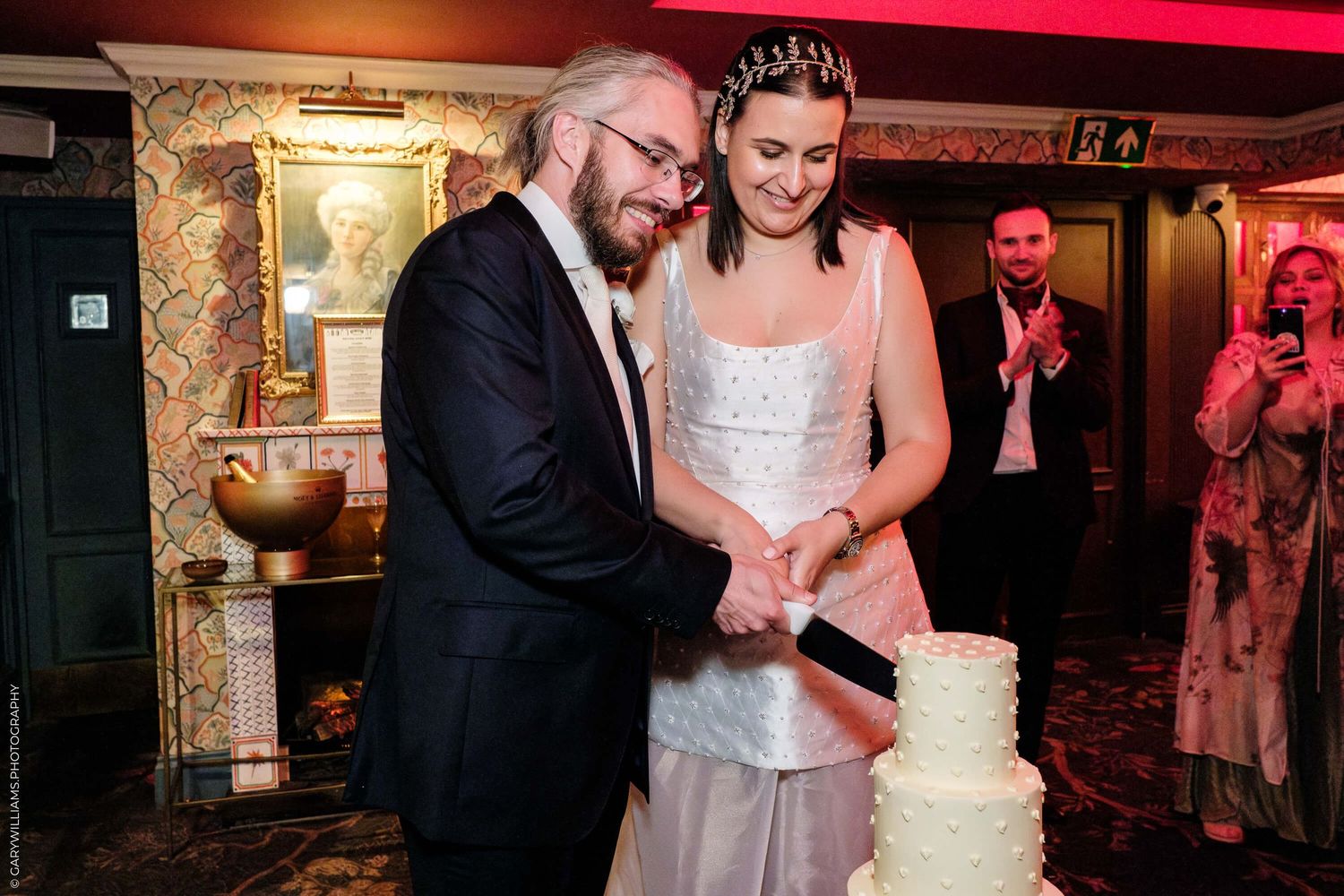 A couple cuts into their white tiered wedding cake together during reception at candlelit venue.