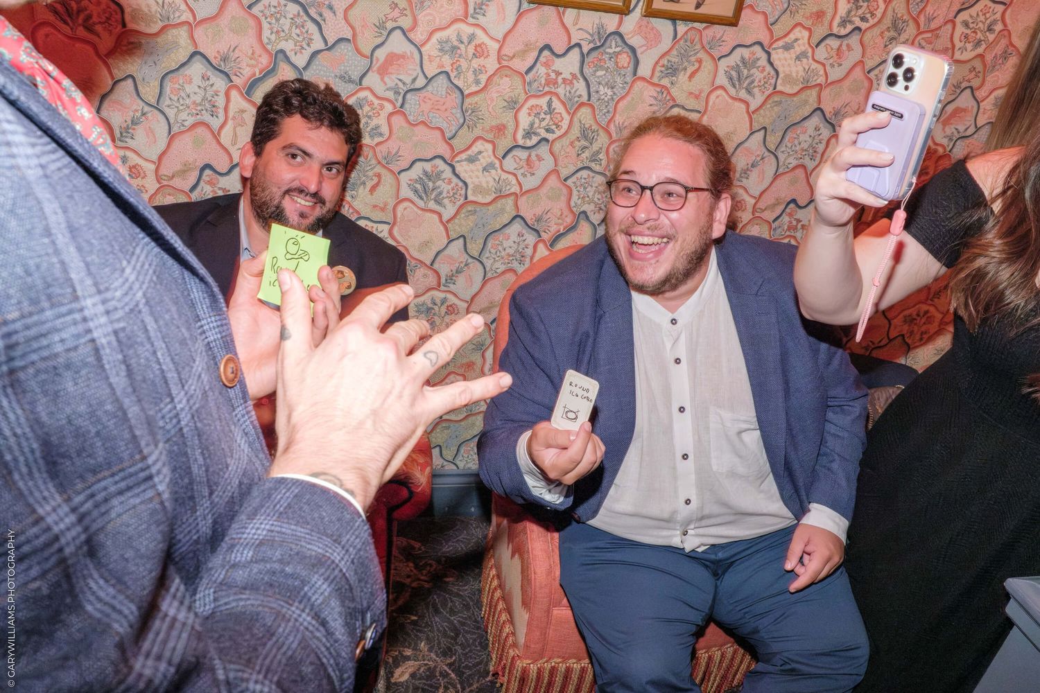 People sitting together enjoying drinks and conversations at a casual social gathering with a pink wallpapered background.