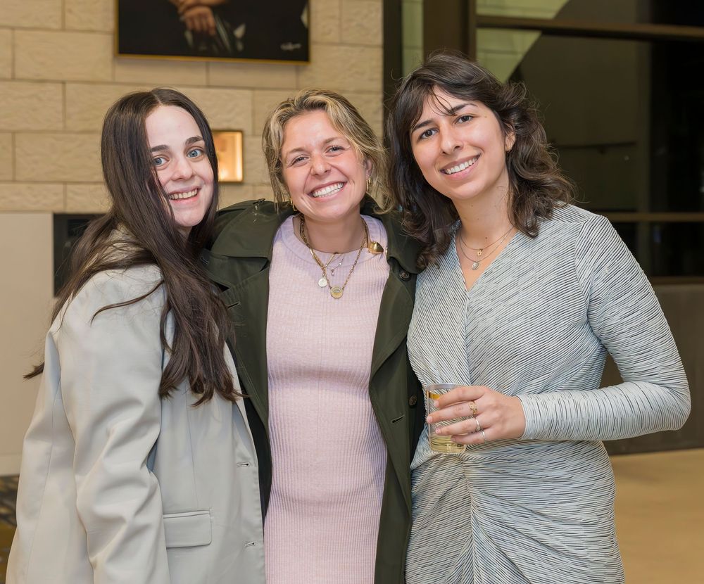Three people in casual attire standing together and smiling during an indoor gathering.