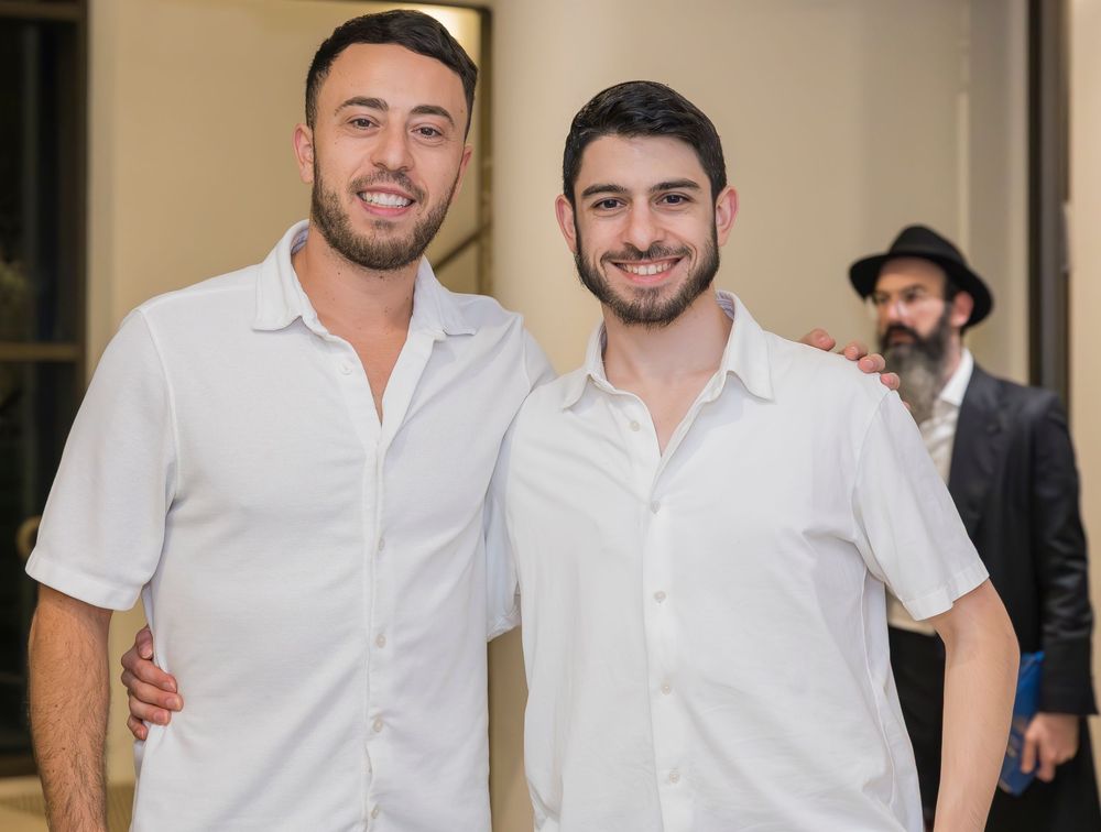 Two people in white polo shirts smiling together with someone wearing a hat visible in the background.
