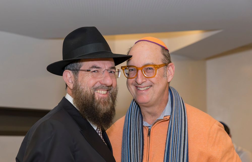 Two people sharing a friendly moment indoors, one wearing a black hat and beard, the other in orange with glasses.