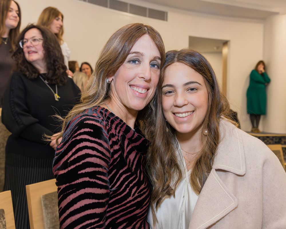 Two people smiling together at an indoor gathering, one in a striped top and one in a light coat.