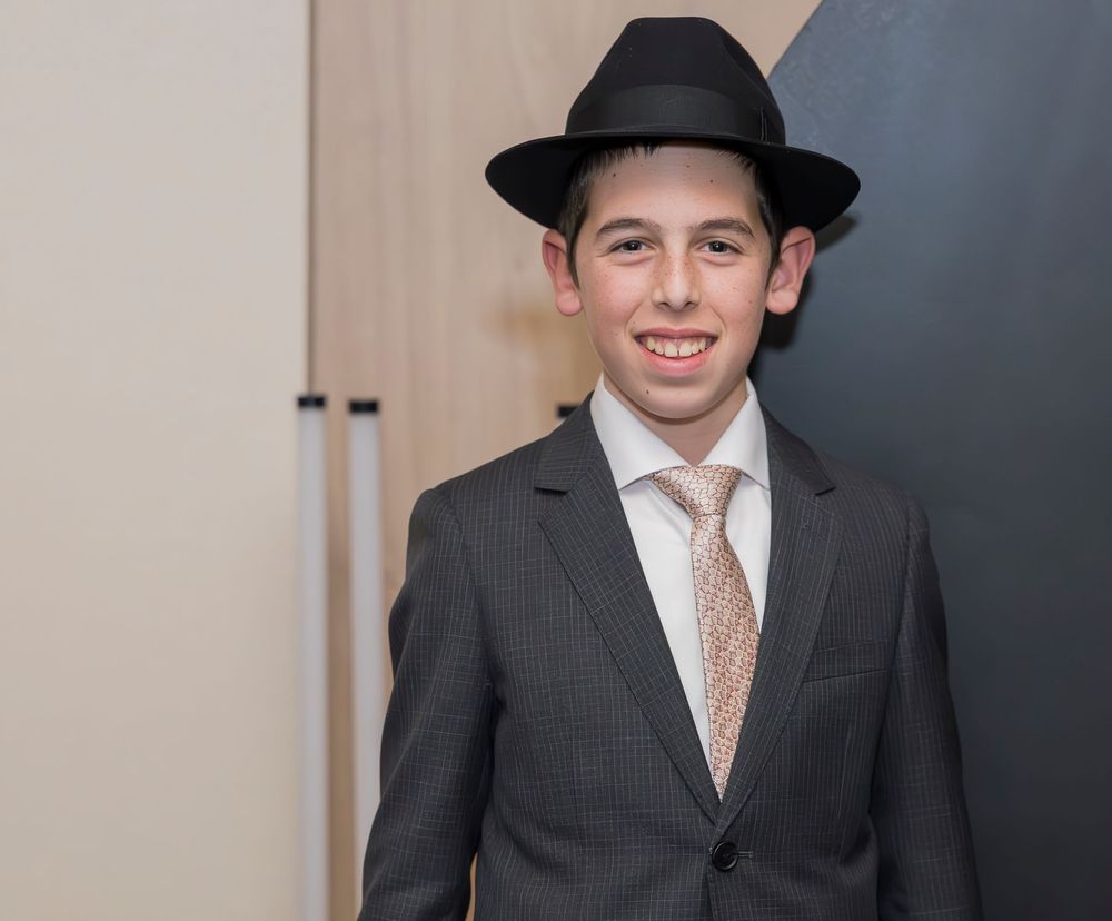 Young person in a formal dark suit and hat poses confidently against a light background.