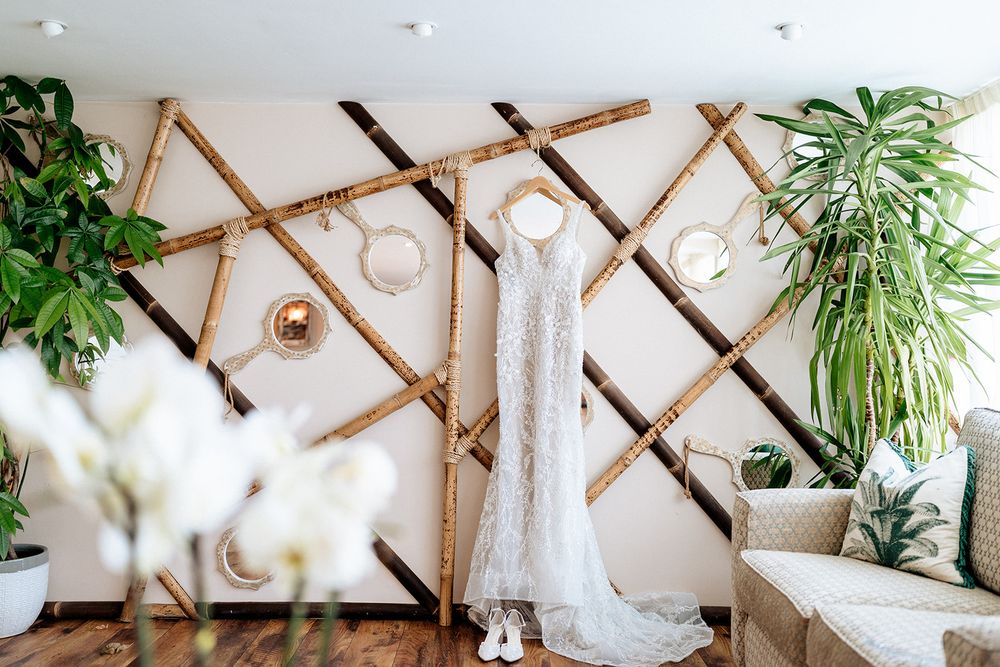 A delicate white lace wedding dress hangs on a bamboo wall display with potted plants and decorative details.