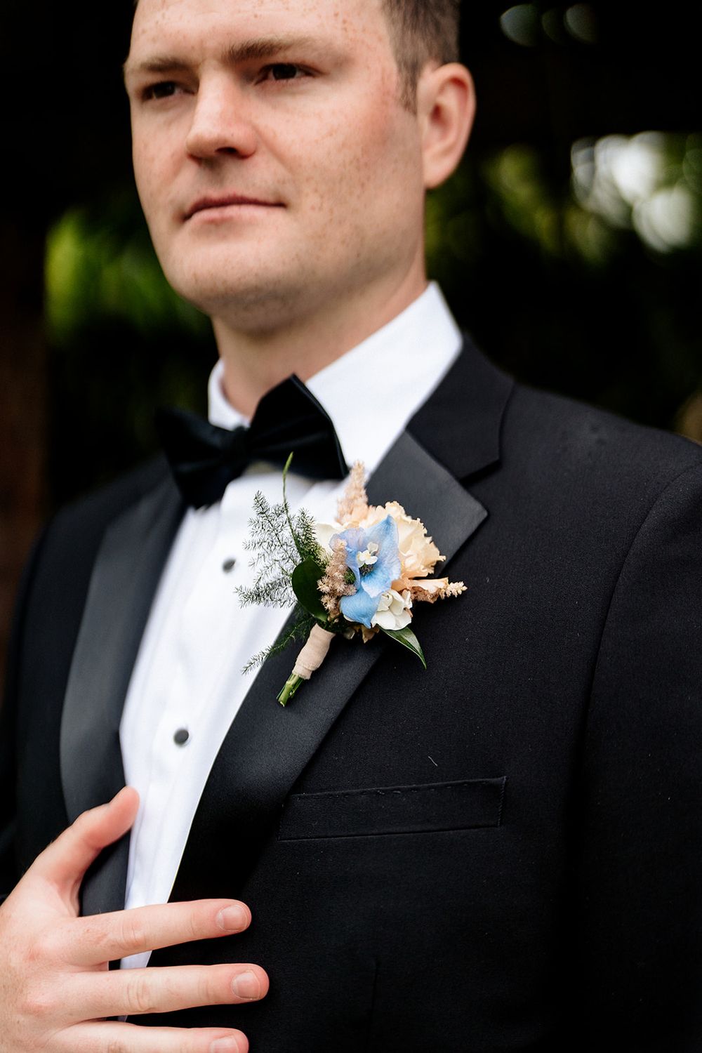 Close-up view of a groom in a black tuxedo and bow tie with a delicate blue and white floral boutonniere.
