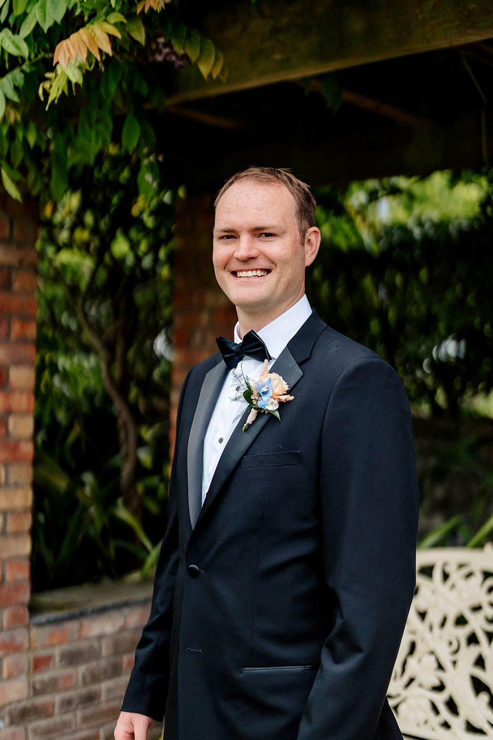A smiling gentleman in a formal black tuxedo with a boutonniere stands under a gazebo with greenery.