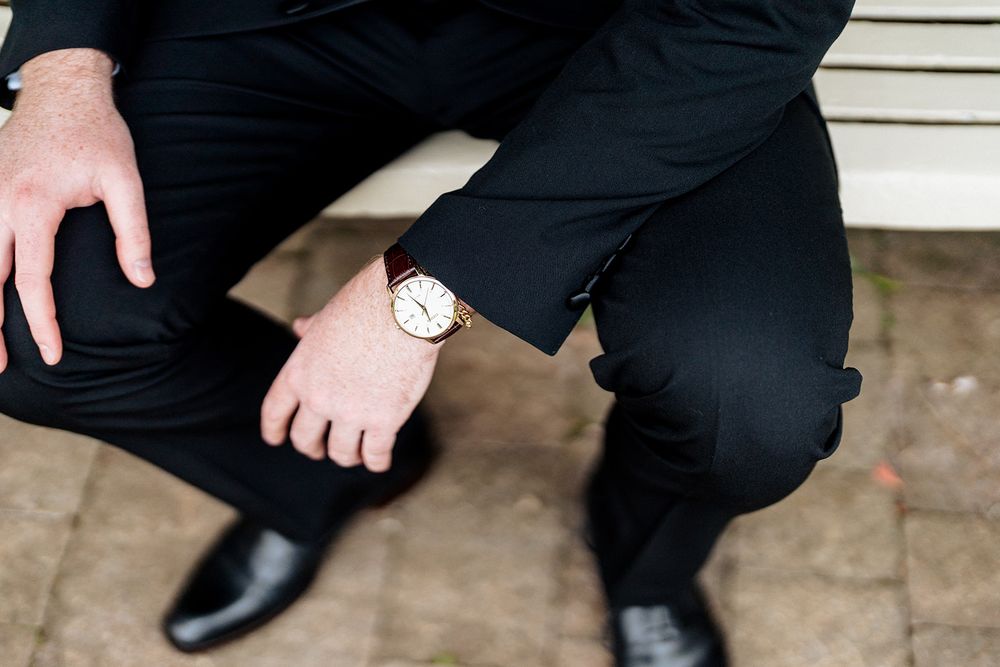Male figure stands against white siding while polishing black leather dress shoes with shoeshine brush.