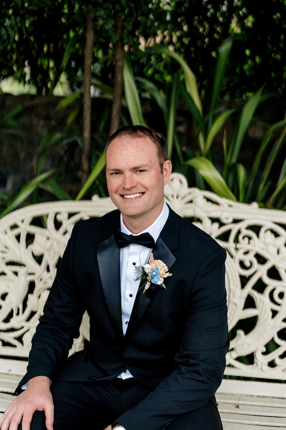 A groom in a black tuxedo and boutonniere sits on an ornate white bench with lush tropical foliage in the background.