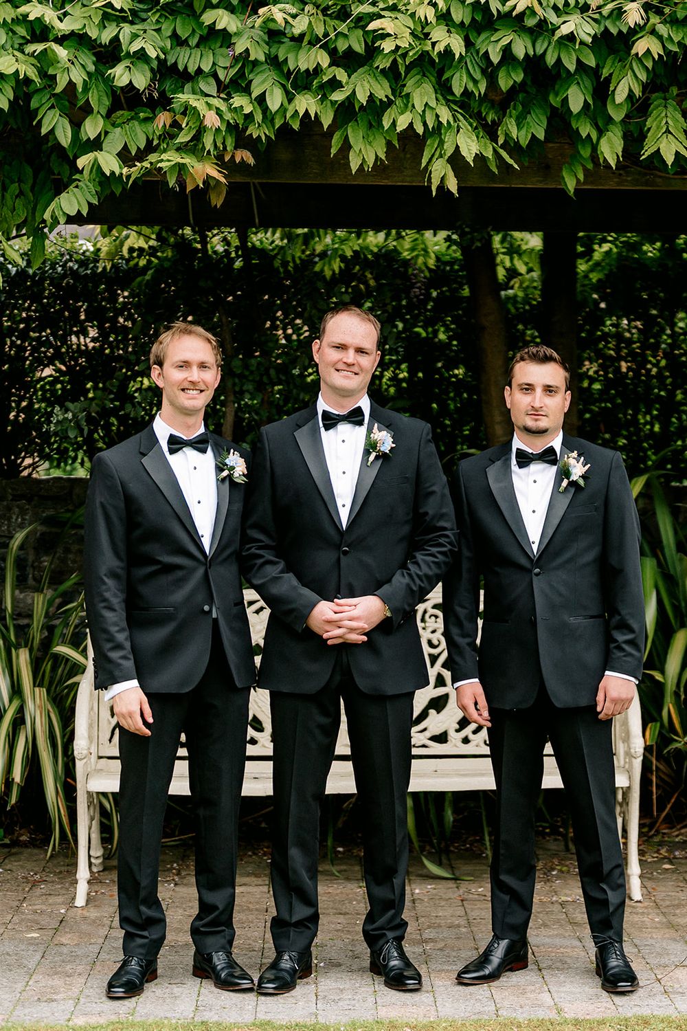 Three groomsmen wearing matching black tuxedos and bow ties stand together in front of a garden bench.