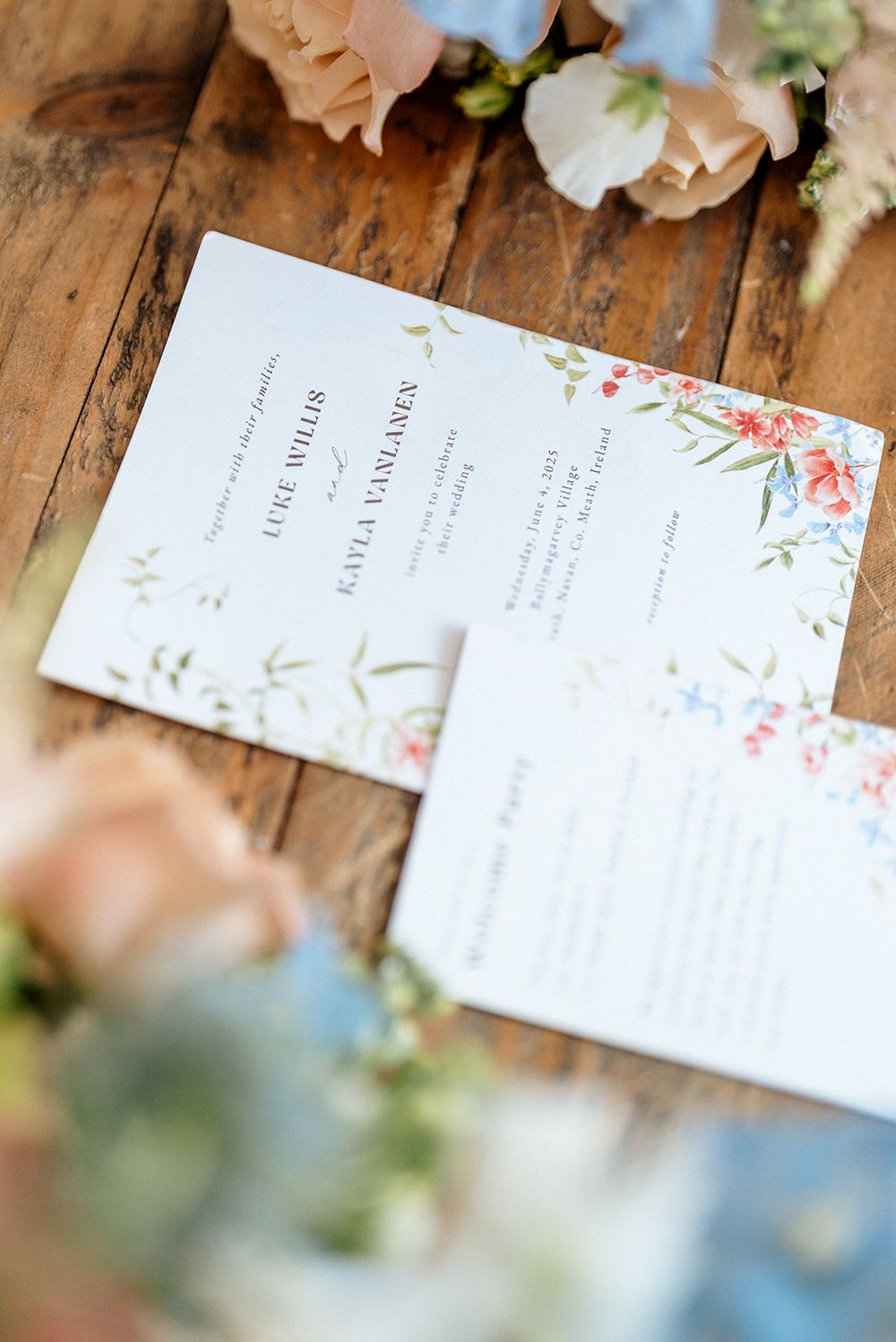 Wedding invitation card with delicate floral watercolor design on a rustic wooden surface with soft florals in background.