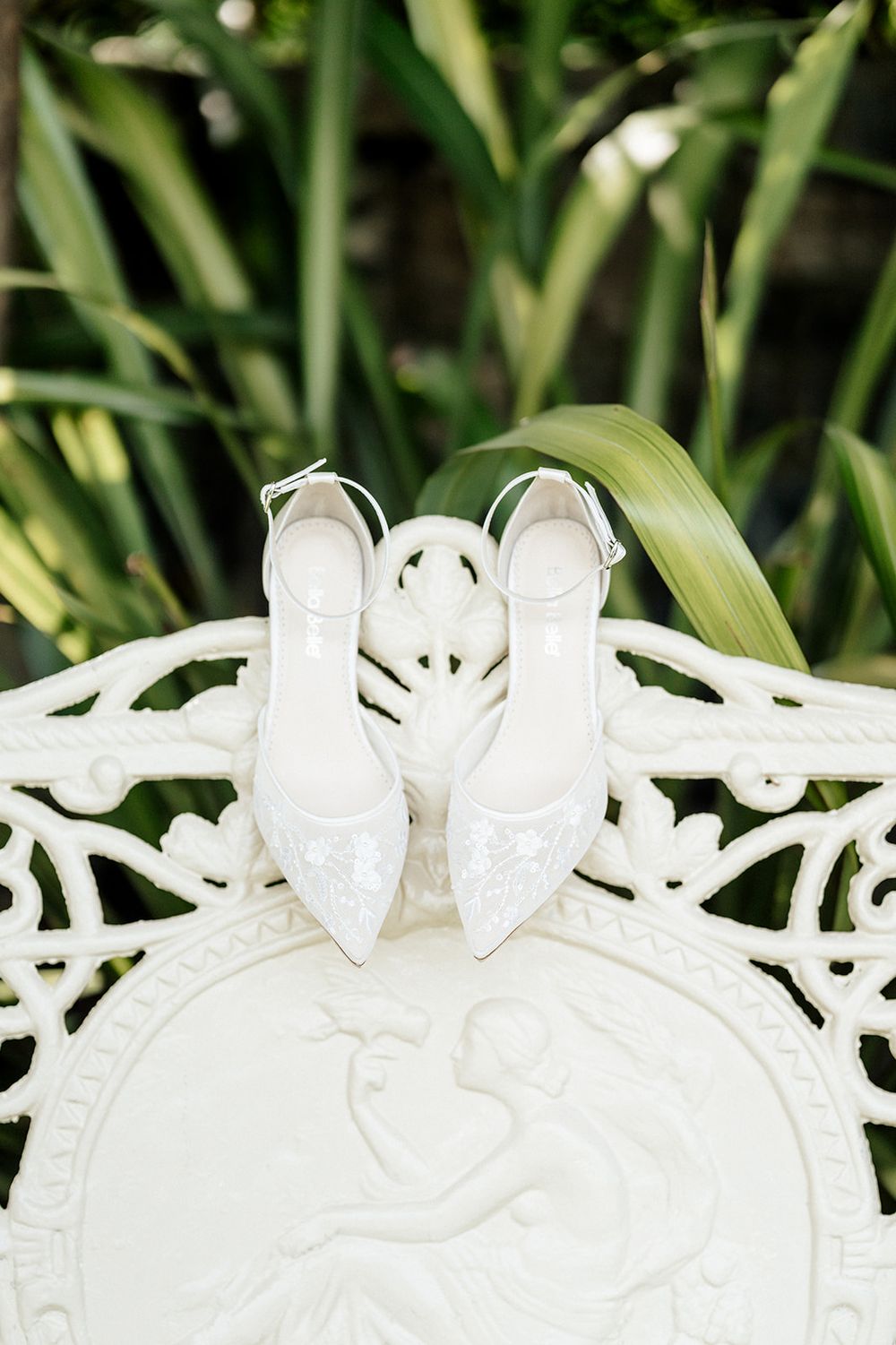 Elegant white bridal shoes displayed on ornate white ironwork furniture with green foliage in the background.