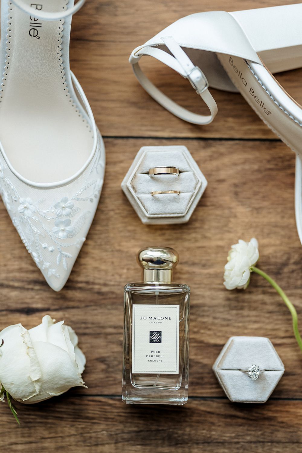 Elegant white bridal shoes with lace details alongside perfume bottle, wedding rings in hexagonal boxes and white roses.
