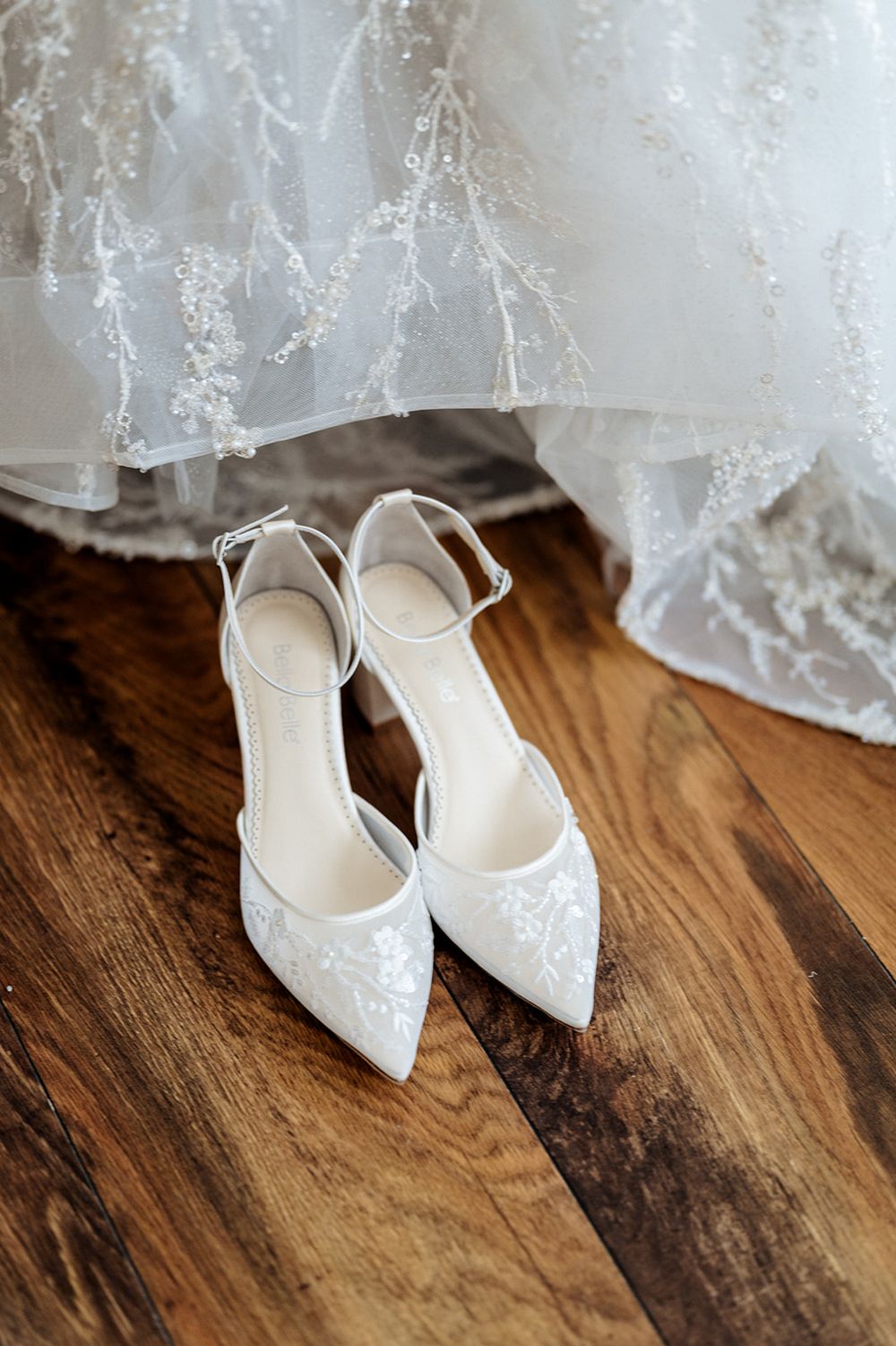 Elegant white lace wedding heels sit on a wooden floor beside delicate tulle fabric.