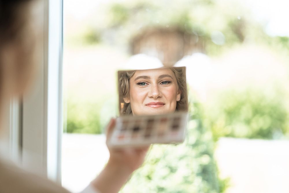 A blurred reflection appears in a handheld mirror with soft green foliage visible in the background.