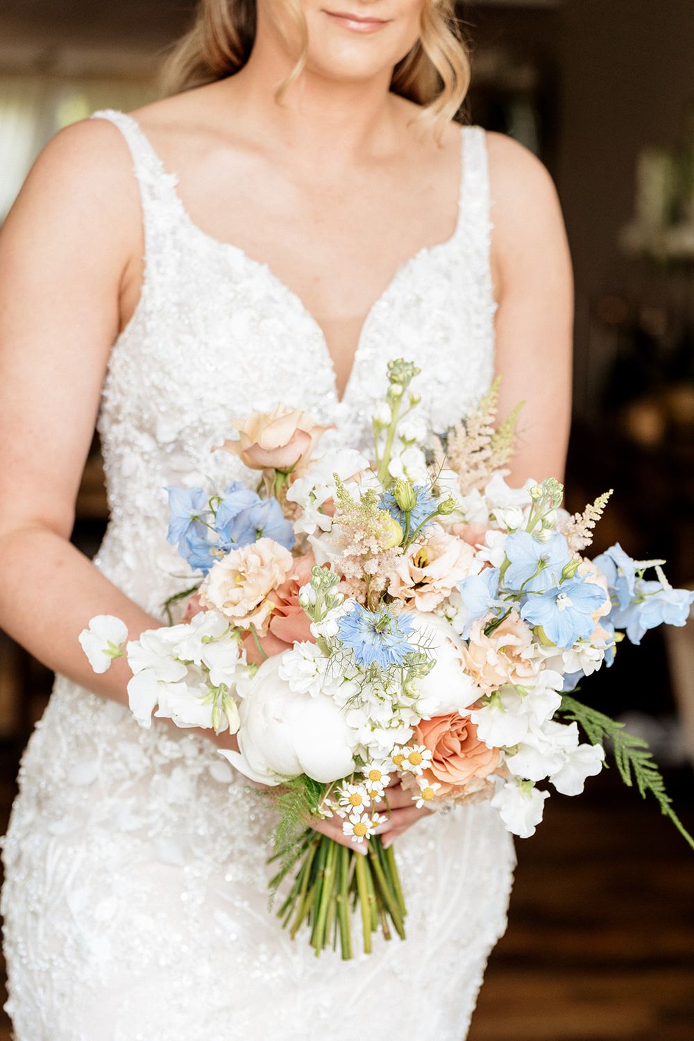 A bridal bouquet featuring delicate blue, peach and white flowers held against a lace wedding dress.