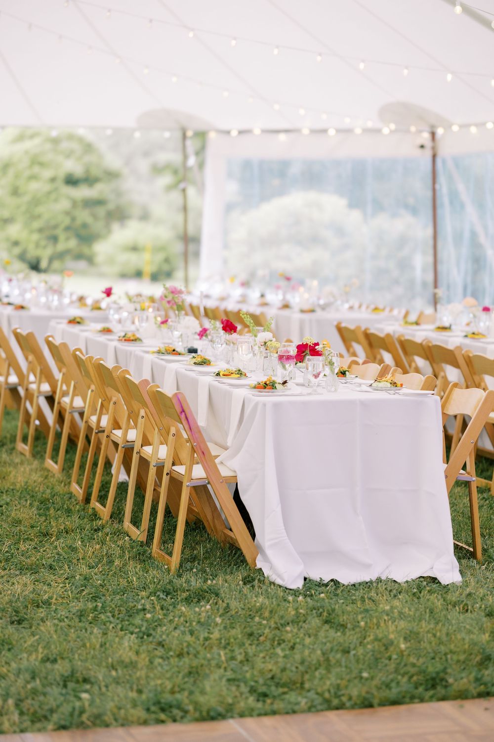 Long banquet tables with white linens and wooden chairs set up under an elegant outdoor tent for a wedding reception.